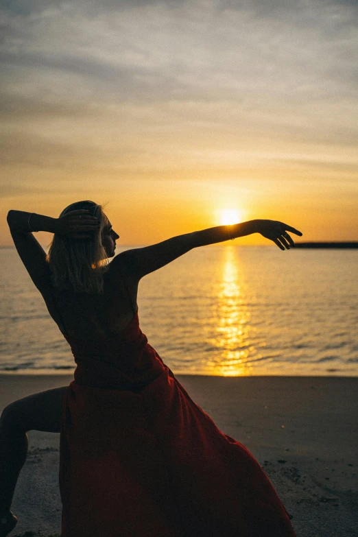 a woman in a red dress on the beach at sunset, pexels contest winner, arabesque, yoga pose, arms out, low sun, doing a sassy pose