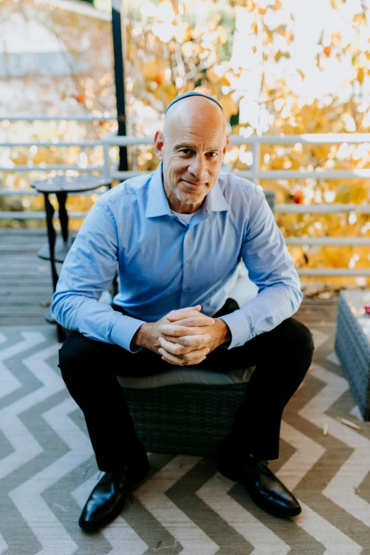 a man in a blue shirt and black pants sitting on a bench, bald head, david lazar, at home, vp of marketing