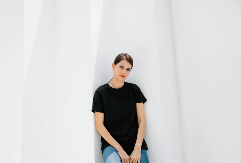 a woman in a black shirt and jeans leaning against a white wall, a portrait, by Emma Andijewska, pexels contest winner, bjarke ingels, alexandra daddario, monument, avatar image