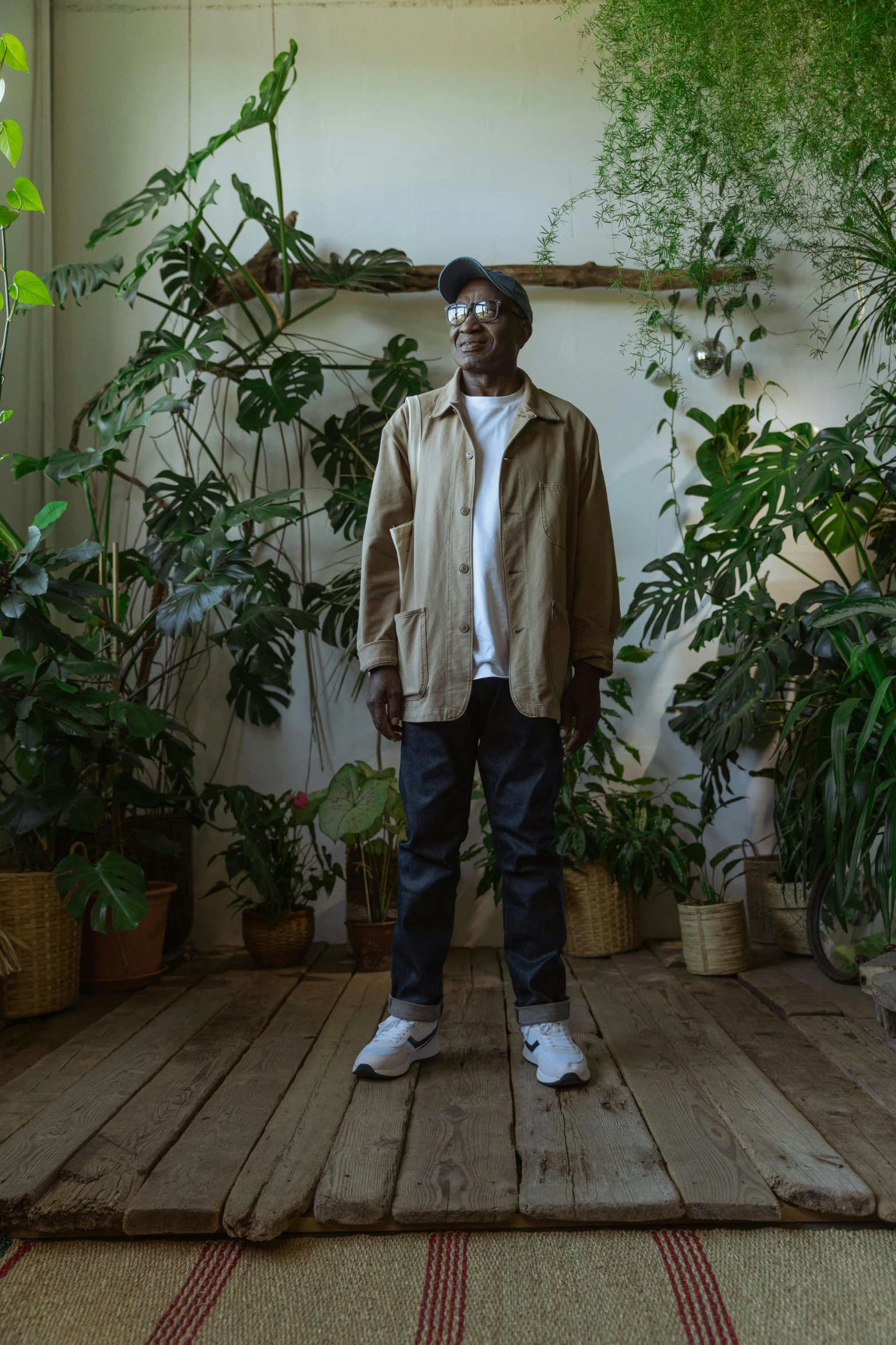 a man standing in a room filled with potted plants, an album cover, inspired by Afewerk Tekle, unsplash, wearing a linen shirt, he is wearing a trenchcoat, blind brown man, lush surroundings