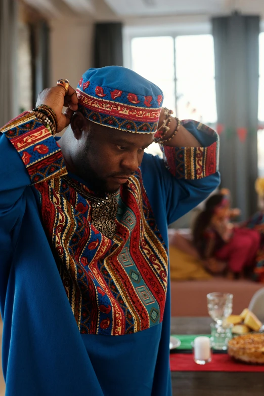 a man standing in a living room next to a table, an album cover, inspired by Oluf Høst, hurufiyya, red and blue garments, ornate turban, 4 k film still, [ theatrical ]