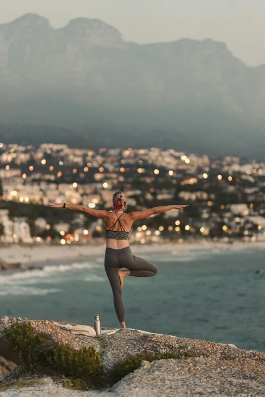 a woman standing on top of a rock next to the ocean, yoga, city views, lit up, south african coast