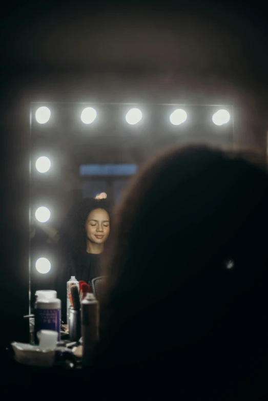 a woman getting her hair done in front of a mirror, by Adam Marczyński, pexels contest winner, ( ( stage lights ) ), theater dressing room, looking straight into camera, panoramic view of girl