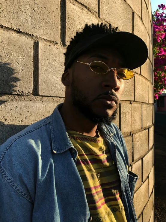 a man standing in front of a brick wall, an album cover, trending on unsplash, photorealism, yellow carrera glasses, refracted sunset lighting, playboi carti portrait, wearing sunglasses and a cap