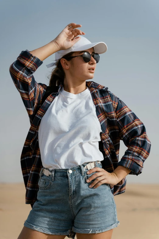 a woman standing on top of a sandy beach, trending on pexels, renaissance, plaid shirt, white baseball hat, an aviator jacket and jorts, young middle eastern woman