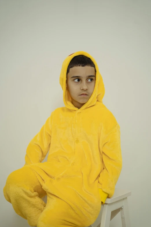 a young boy in a yellow onesuit sitting on a stool, an album cover, pexels, hyperrealism, hooded, hasan piker, wearing pajamas, upset