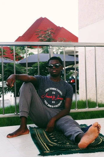 a man sitting on a rug on a balcony, by Chinwe Chukwuogo-Roy, with a cool pose, low quality photo, tourist photo, slightly pixelated
