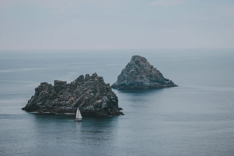 a sailboat in the middle of a large body of water, a picture, by Adam Marczyński, unsplash contest winner, two medium sized islands, jagged rocks, grey, views to the ocean