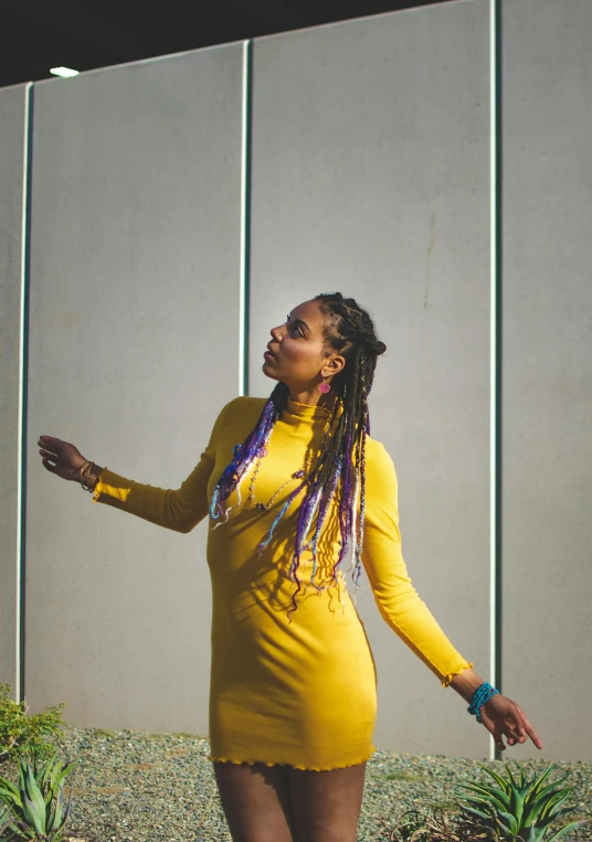 a woman in a yellow dress standing in front of a wall, pexels contest winner, black arts movement, long dreadlocks, yellow and purple color scheme, looking upward, bay area