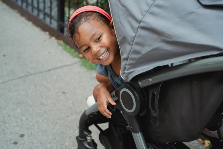 a little girl that is sitting in a stroller, by Jason Felix, smiling into the camera, close up shot from the side, high angle shot, on sidewalk