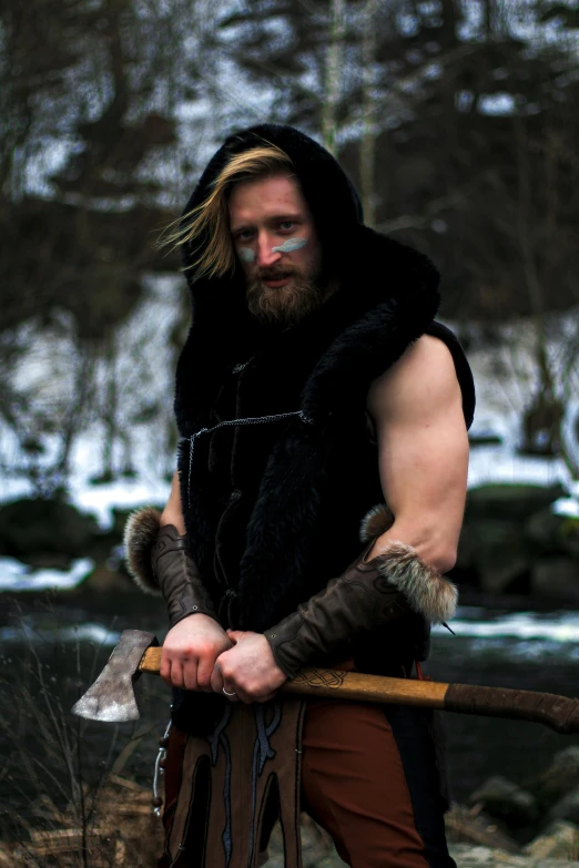 a man standing next to a river holding an axe, inspired by Jóhannes Sveinsson Kjarval, reddit, wearing barbarian caveman pelt, dressed in black body armour, only snow in the background, blond beard