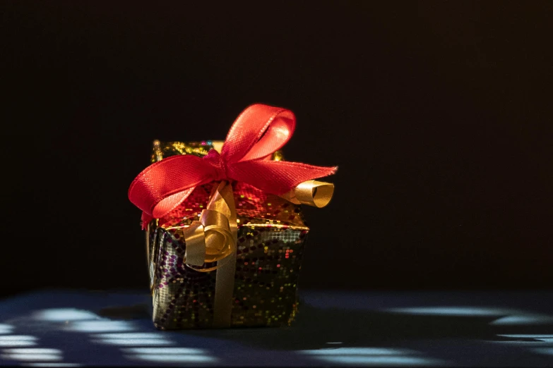 a gift box with a red bow sitting on a table, by Julia Pishtar, pexels contest winner, photorealism, shining gold and black and red, red and blue back light, close up shot of an amulet, snacks