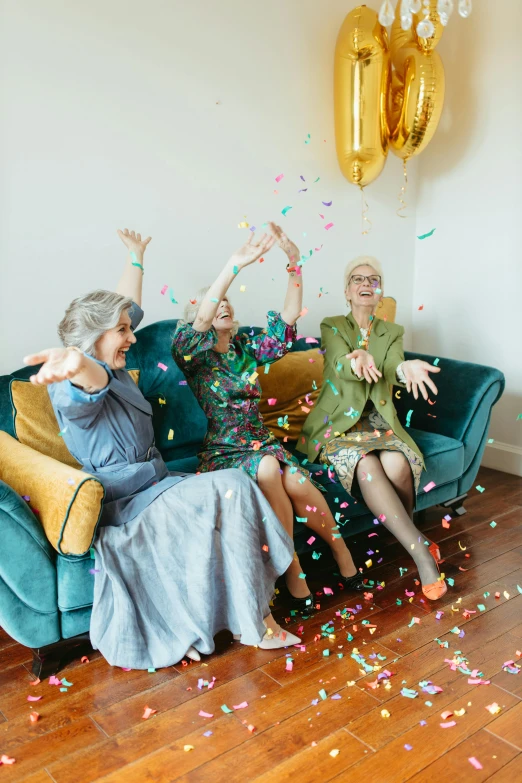 a group of people sitting on top of a couch covered in confetti, three women, 70 years old, gold and teal color scheme, non-binary