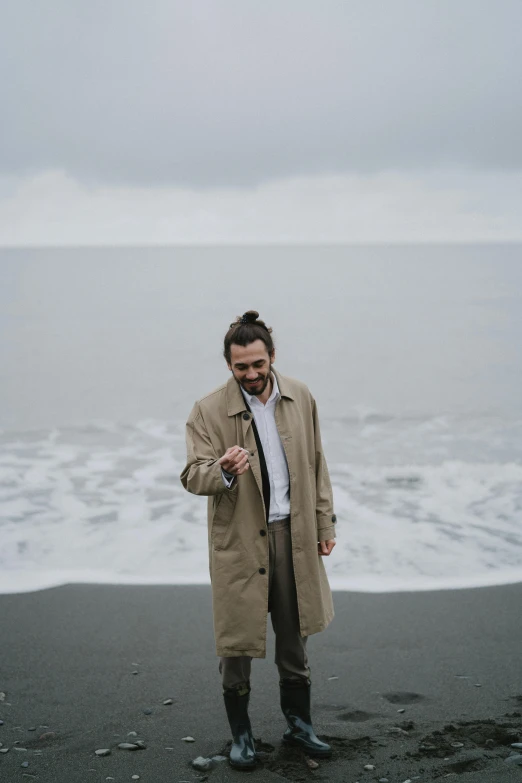 a man standing on top of a beach next to the ocean, light brown trenchcoat, matt berry, on a gray background, jovana rikalo