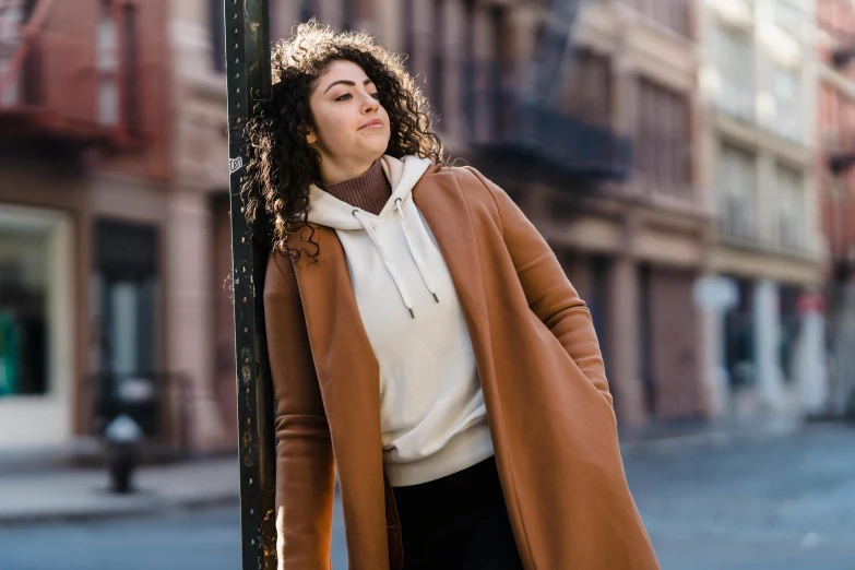 a woman leaning against a pole on a city street, trending on pexels, renaissance, beige hoodie, brown curly hair, brown tuffle coat, young middle eastern woman