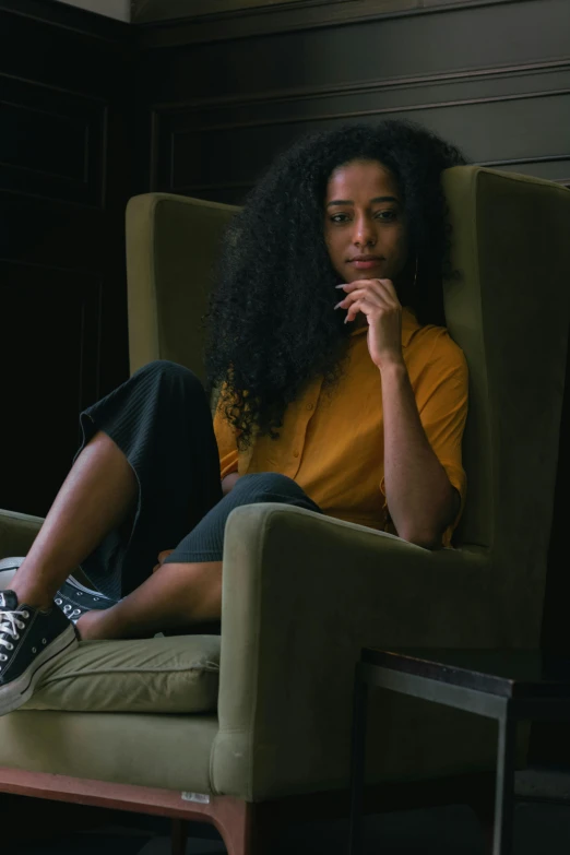 a woman sitting on top of a green chair, by Lily Delissa Joseph, pexels contest winner, black curly hair, wearing casual clothing, black and yellow, androgynous person