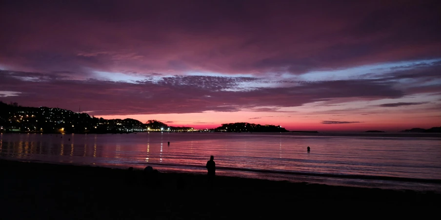 a couple of people standing on top of a beach next to the ocean, barely lit warm violet red light, manly, craigville, from the distance