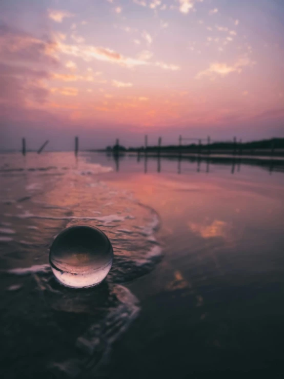 a glass ball sitting on top of a sandy beach, in water, during a sunset, body of water, landscape photograph