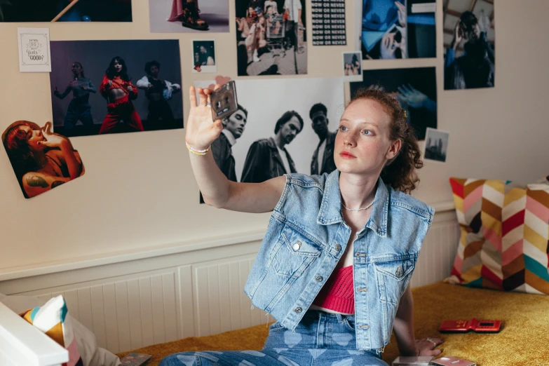 a woman sitting on a bed holding up a cell phone, an album cover, inspired by Nan Goldin, trending on pexels, wearing a jeans jackets, sadie sink, in her art room, wearing a crop top