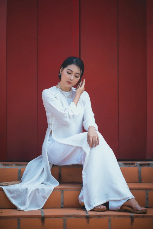 a woman sitting on some steps talking on a cell phone, inspired by Gao Kegong, pexels contest winner, white silky outfit, ao dai, square, thinking pose