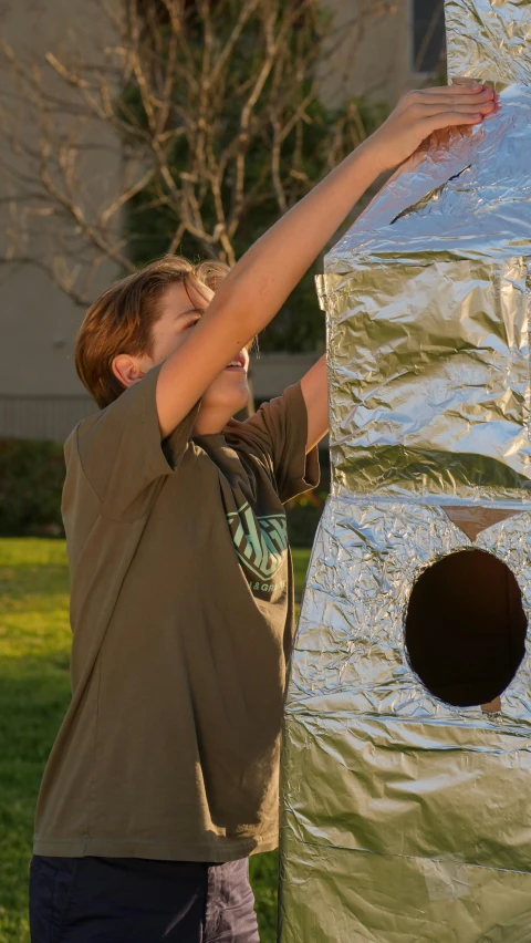 a little boy that is standing in the grass, by Thomas Furlong, interactive art, solar sail in space, made out of shiny silver, cardboard tunnels, holding a tower shield