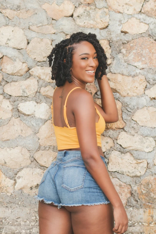 a woman standing in front of a stone wall, by Lily Delissa Joseph, trending on unsplash, wearing yellow croptop, curls hair, jamaica, arched back