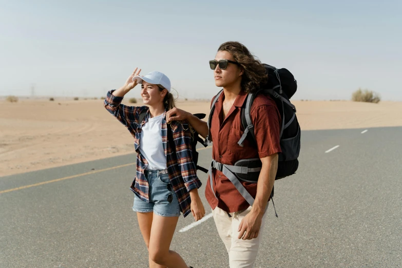 a man and a woman standing on the side of a road, trending on pexels, a man wearing a backpack, in the desert beside the gulf, college students, baggy clothing and hat