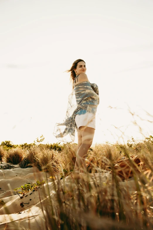 a woman standing on top of a sandy beach, in a grass field, maternity feeling, long hair shawl, modelling