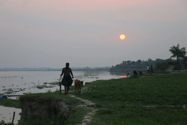 a man riding a bike down a dirt road next to a body of water, by Sunil Das, pexels contest winner, bengal school of art, sunset in the distance, during a blood moon, she is walking on a river, the sun shines in