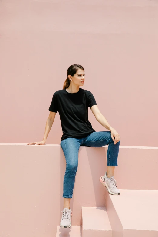 a woman sitting on top of a pink wall, wearing black tshirt, blue jeans and grey sneakers, in front of white back drop, looking off to the side