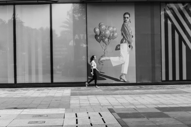 a black and white photo of a woman walking down a sidewalk, a black and white photo, street art, appearing in a shopping mall, balloon, clear glass wall, stylish pose