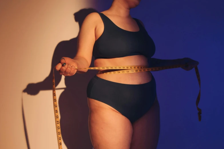 a woman measuring her waist with a tape, by Emma Andijewska, trending on pexels, figuration libre, a portrait of a plump woman, neoprene, dimly - lit, blue undergarments