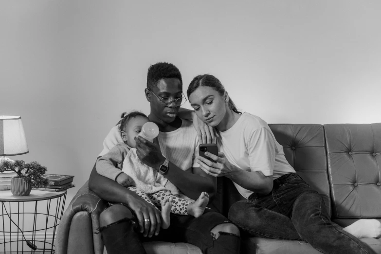a man and woman sitting on a couch with a baby, a black and white photo, pexels, realism, cell phones, black man, lesbians, bella poarch