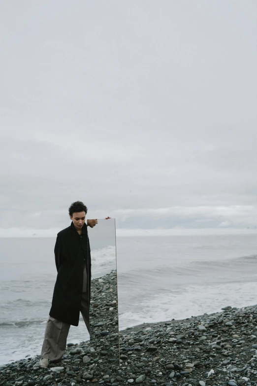 a man standing on a rocky beach next to the ocean, standing in front of a mirror, grey skies, wearing black overcoat, dylan kowalsk