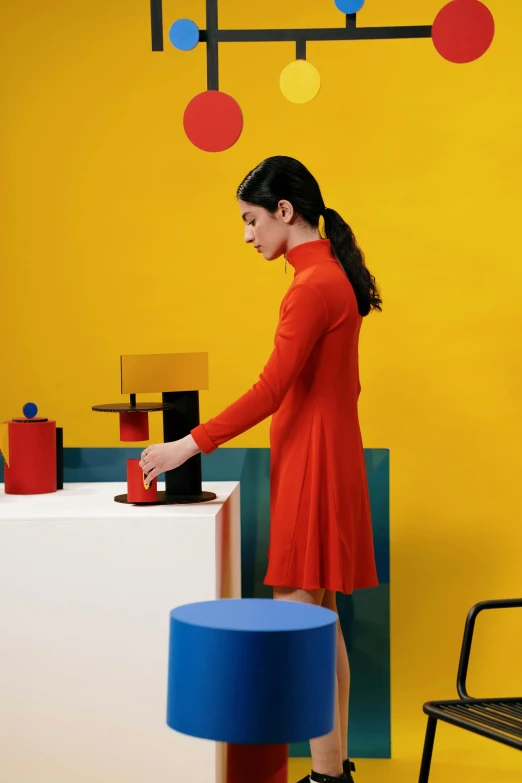 a woman is standing in a brightly colored room, inspired by Bauhaus, product display, red+yellow colours, hand on table, dress