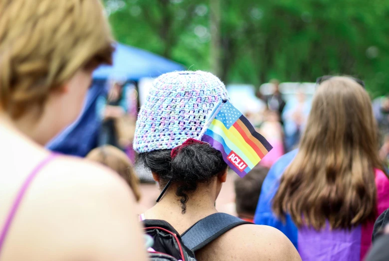 a woman with a hat that has a rainbow flag on it, a photo, audience in the background, back of head, park in background, covered head