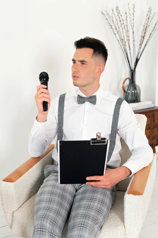a man sitting in a chair holding a microphone and a clipboard, inspired by Oskar Lüthy, featured on reddit, romanticism, well-groomed model, platinum attire, official product image, bowtie