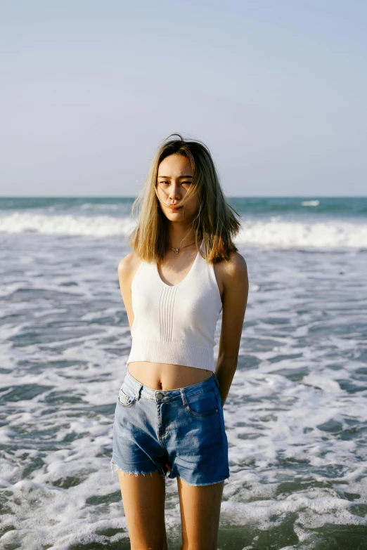 a woman standing on top of a beach next to the ocean, wearing : tanktop, kiko mizuhara, white shirt and jeans, wavy hair