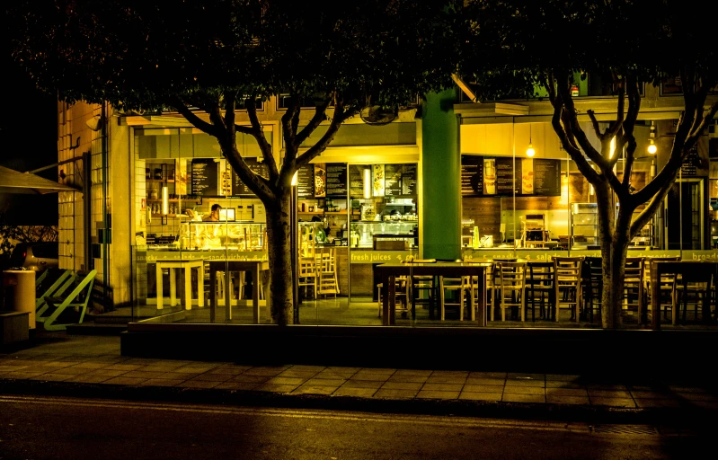 the outside of a restaurant lit up at night, by Lee Loughridge, unsplash, green square, bakery, sitting down, in a row