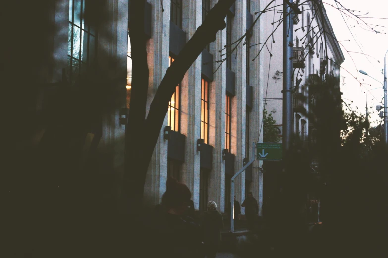 a group of people walking down a street next to tall buildings, a photo, inspired by Elsa Bleda, modernism, massive trees with warm windows, 2 0 0 0's photo, sundown, old library