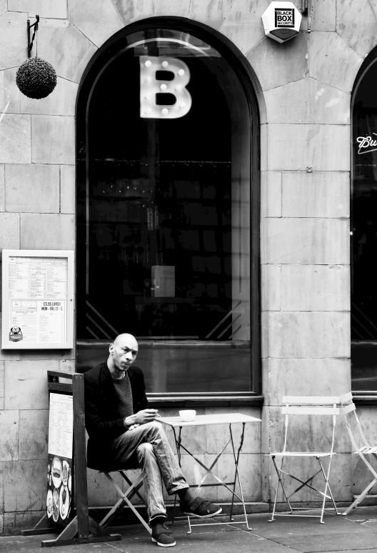 a man sitting on a chair in front of a building, a black and white photo, cafe, babel, bald man, in barcelona
