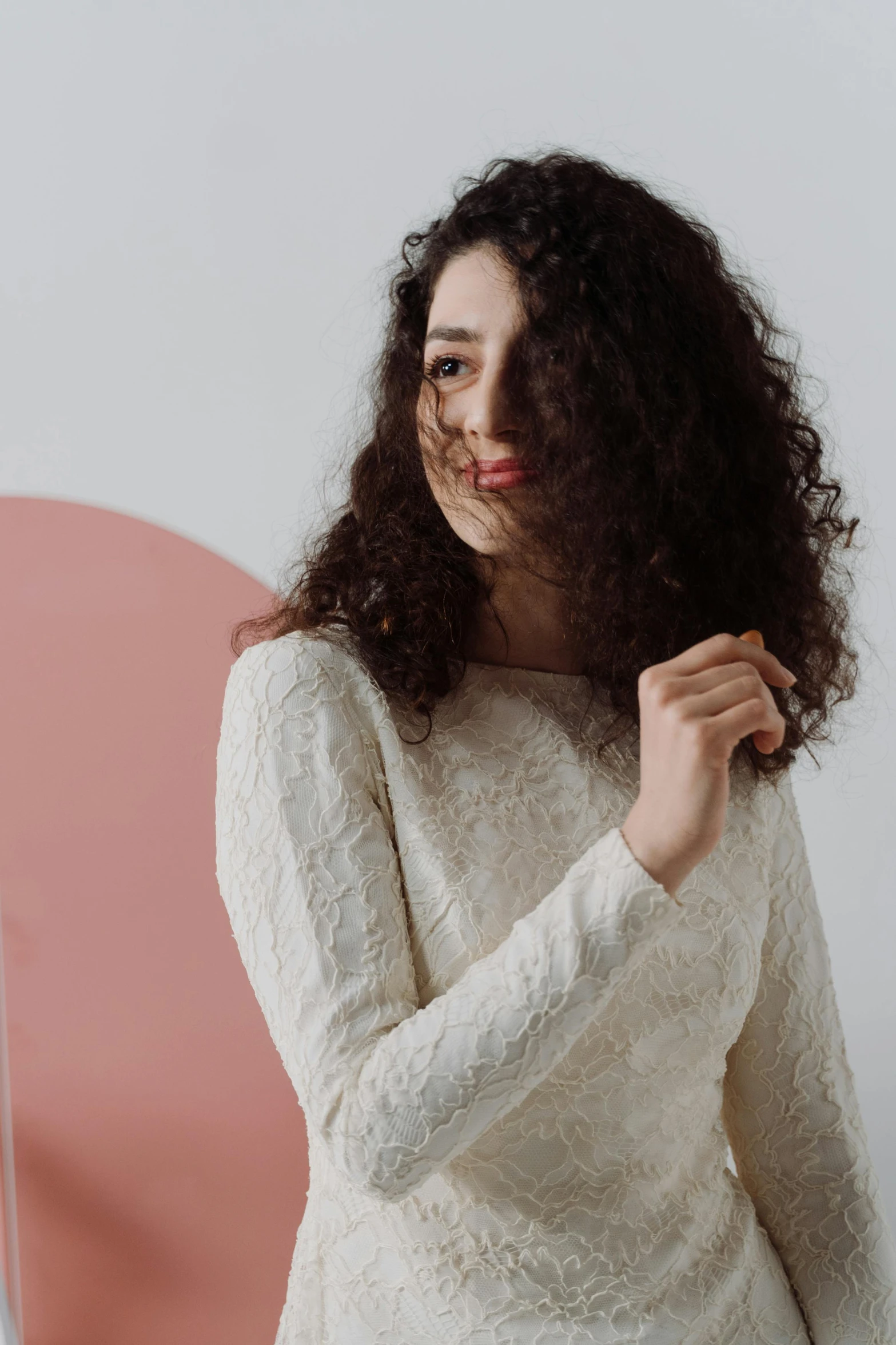 a woman standing in front of a pink wall, curly dark hair, wearing a white sweater, in white room, pale-skinned persian girl