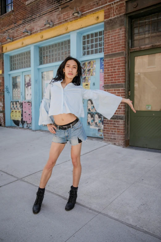 a woman standing on a sidewalk in front of a building, croptop and shorts, wearing a light blue shirt, posing like a falling model, yael shelbia