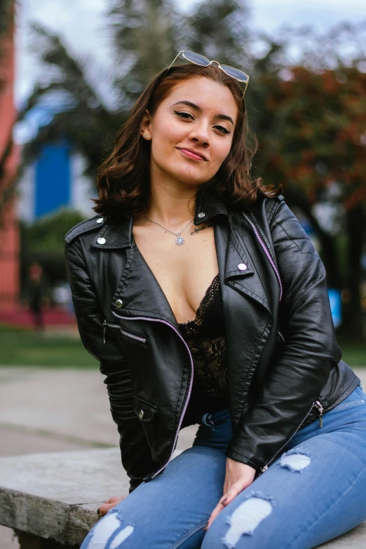 a woman sitting on top of a cement bench, wearing a black leather jacket, wearing bra, chilean, profile image