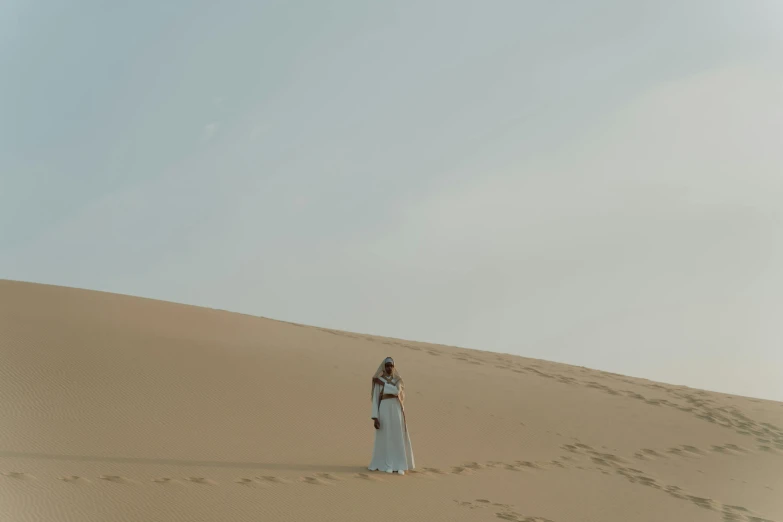 a woman standing in the middle of a desert, an album cover, pexels contest winner, in a long white dress, blue, sand color, low angle wide shot