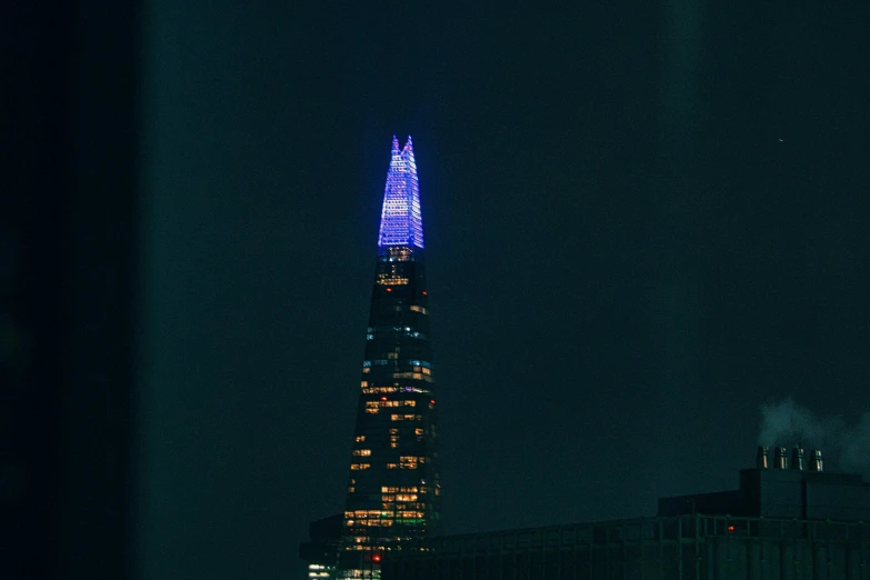 a very tall building lit up at night, spire, purple and blue leather, serrated point, blue and orange