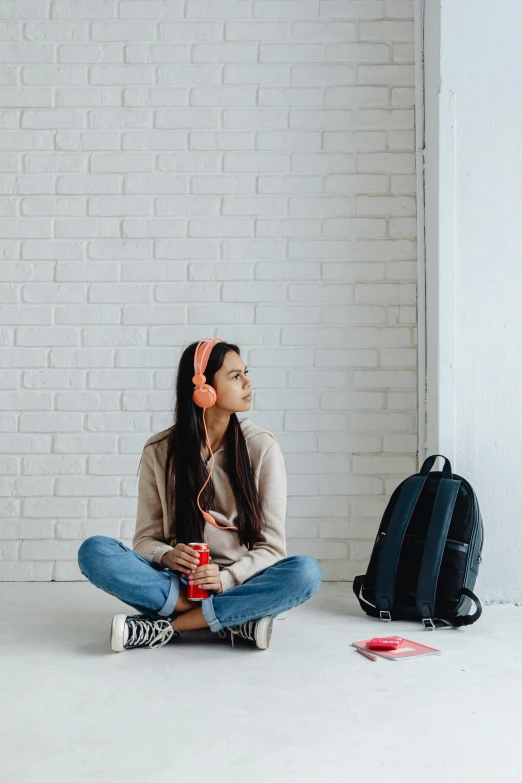 a woman sitting on the floor in front of a brick wall, trending on pexels, girl wearing headphones, sitting in the classroom, pondering, with a backpack