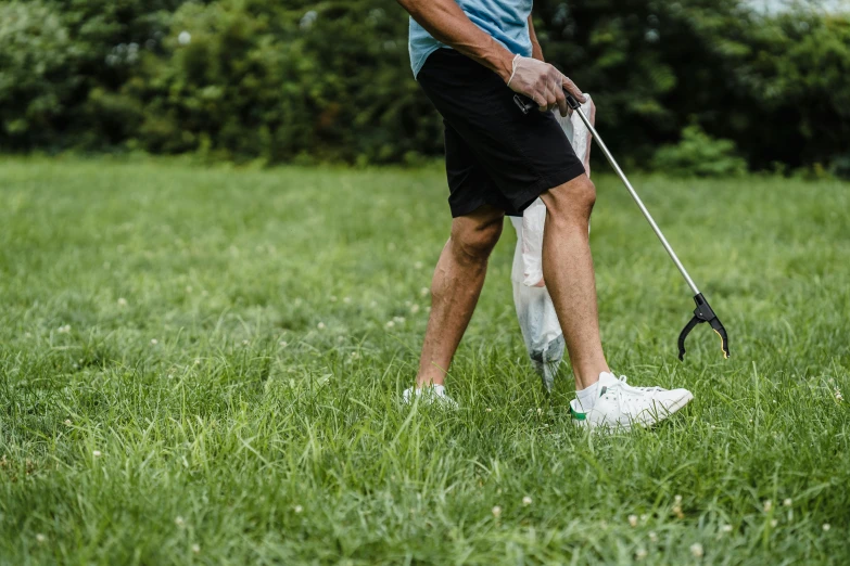 a man standing on top of a lush green field, crutches, wearing golf shorts, old man doing with mask, using a spade