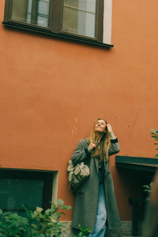 a woman standing in front of a building talking on a cell phone, by Anna Boch, pexels contest winner, light brown coat, looking upwards, with a backpack, desaturated color