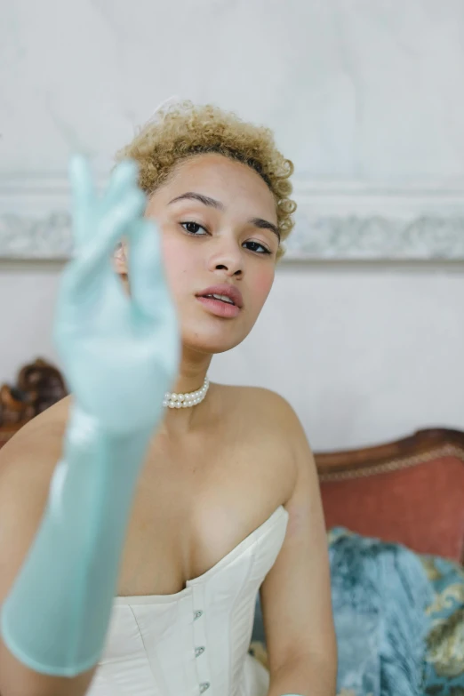 a woman in a white dress and blue gloves, inspired by Cecil Beaton, trending on pexels, visual art, short blonde afro, pastel green, hand gesture, glossy skin